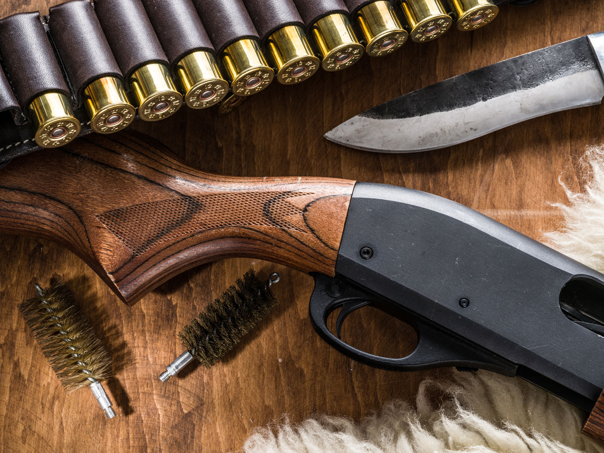 A shotgun, a cartridge belt of shells, a hunting knife, and cleaning brushes laid out on wood